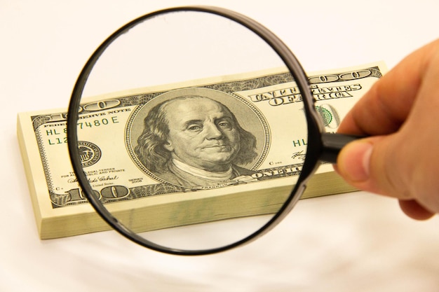 A person holding a magnifying glass over a document with dollar signs on it, symbolizing the detailed search and investigation needed to find unclaimed money.